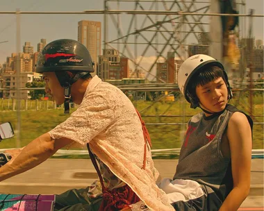 Two people on a motorcycle, wearing helmets, with an urban cityscape in the background.