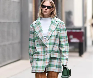 Woman in a plaid blazer, white top, and brown pants, holding a green bag, wearing sunglasses on a city street.