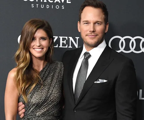 A smiling couple poses on the red carpet; she wears a patterned dress, and he wears a suit and tie, with sponsor logos behind.