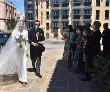 Bride in white gown and veil entering venue, accompanied by man in suit, with masked guests watching.