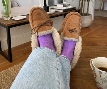 Cozy brown moccasin slippers and purple socks on a person lounging in jeans, with a floral mug on the wooden floor.