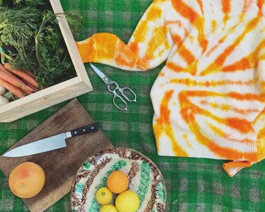 Orange tie-dye sweater on green checkered table with fruit bowl, knife, box of veggies, and scissors.