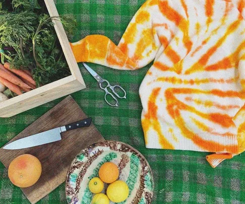 Orange tie-dye sweater on green checkered table with fruit bowl, knife, box of veggies, and scissors.