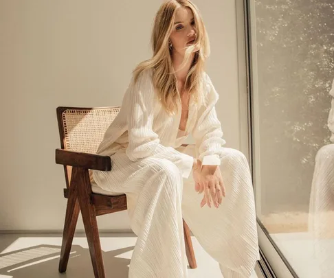 A woman in a flowing white outfit sits on a wooden chair by a sunlit window, with serene expression.