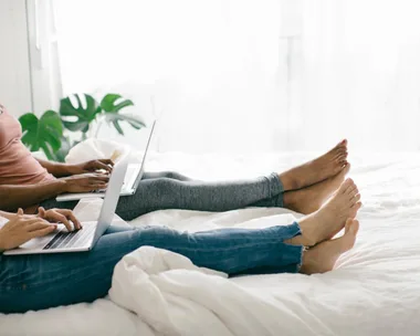 Two people working on laptops while sitting on a bed, legs stretched out, with a plant in the background.