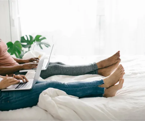 Two people working on laptops while sitting on a bed, legs stretched out, with a plant in the background.