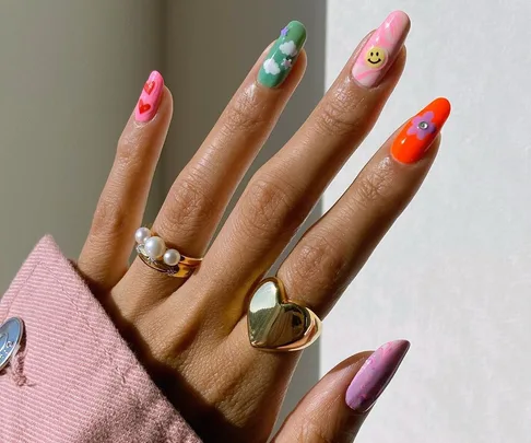 Hand with colorful nail art designs, including smiley face, hearts, and flowers, wearing pearl and heart rings.