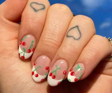 Close-up of a hand with a cherry-themed French manicure and heart tattoos on two fingers.