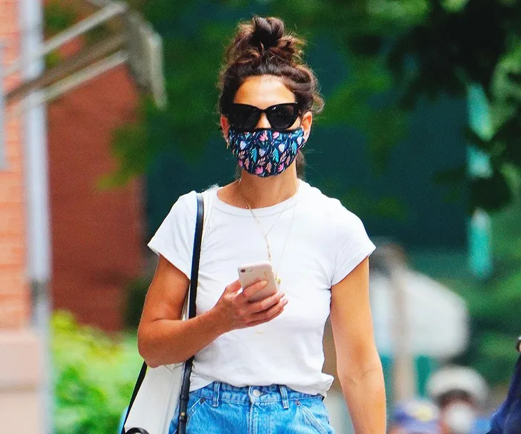 Person wearing a white shirt, jeans, printed face mask, and sunglasses, holding a phone while walking outside.