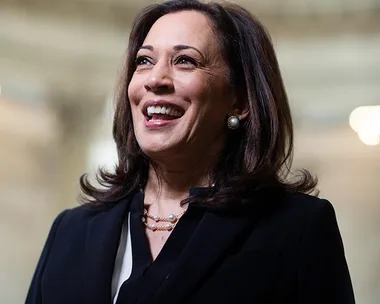 A smiling woman in formal attire with pearl earrings and necklace standing indoors.