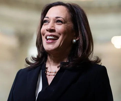 A smiling woman in formal attire with pearl earrings and necklace standing indoors.