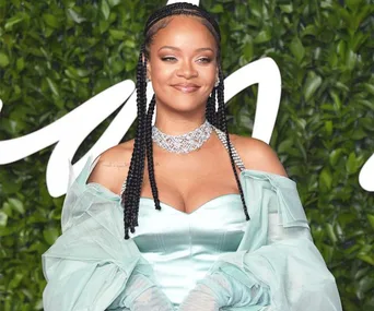 A woman in a light blue dress with braids and a statement necklace, standing in front of a leafy background.