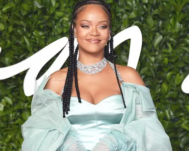 A woman in a light blue dress with braids and a statement necklace, standing in front of a leafy background.