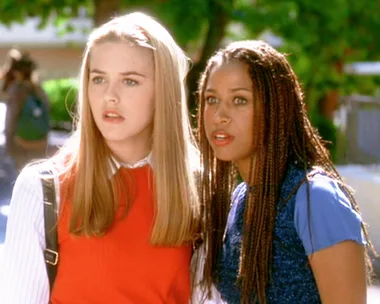 Two teenage girls from "Clueless" look surprised outdoors; one wears a red sweater, and the other a blue top.