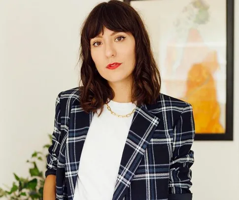 A person with dark hair in a plaid blazer and white shirt stands indoors, with a framed artwork in the background.