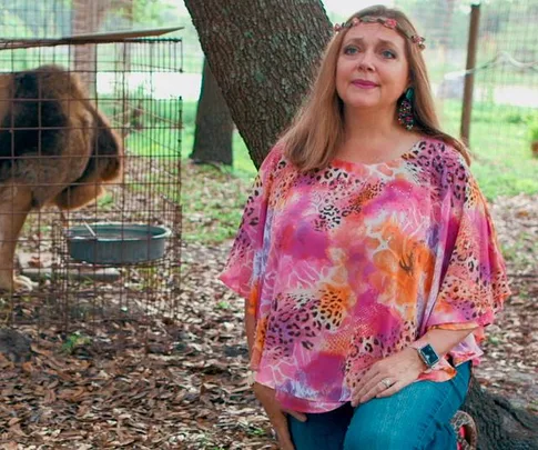 Woman in floral top kneels beside a fenced lion, outdoors in a wooded area.