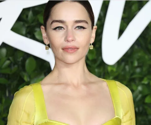 A woman in a yellow dress with dark hair, standing in front of a leafy backdrop at an event.