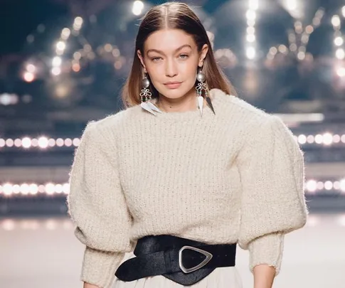 Gigi Hadid walking a fashion runway wearing a cream sweater, large earrings, and a wide black belt.