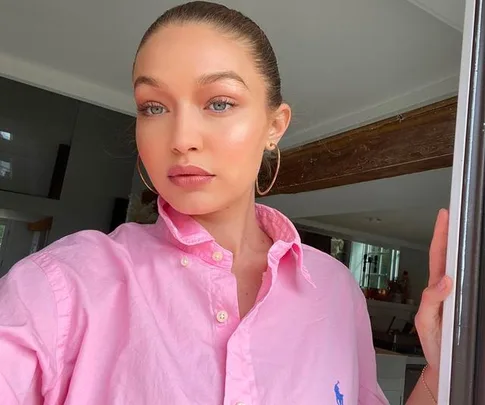 A woman in a pink shirt stands indoors, looking into the camera with natural light illuminating her face.