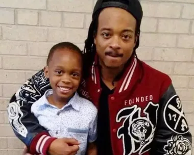 Man with dreadlocks in red jacket and a smiling child in a white shirt hug in front of a brick wall.