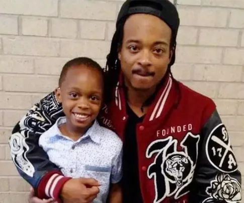 Man with dreadlocks in red jacket and a smiling child in a white shirt hug in front of a brick wall.