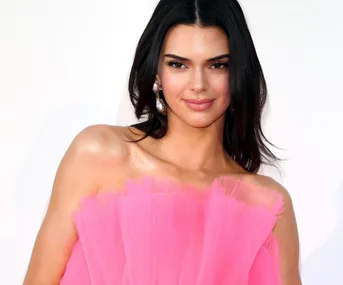 A woman in a pink off-shoulder dress poses with a subtle smile against a plain background.