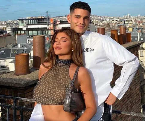 Two people posing on a rooftop with a cityscape backdrop, one wearing a patterned top and holding a small bag.