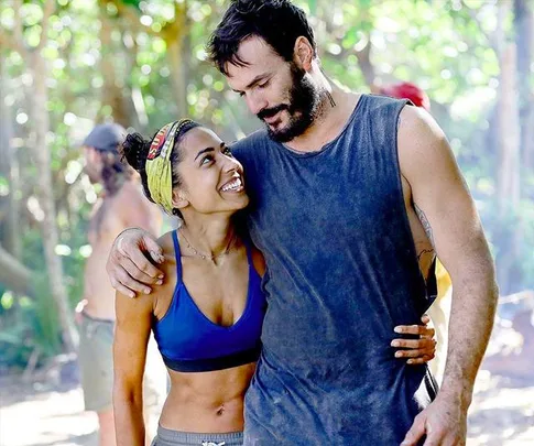 Man and woman smiling at each other outdoors, arms around shoulders, wearing casual sportswear.