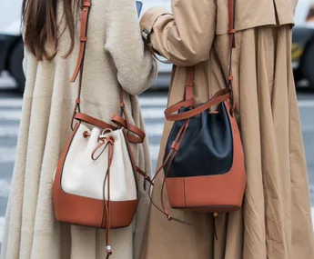 Two women wearing trench coats carrying stylish bucket bags on a city street.