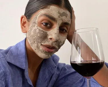 Person with a clay face mask and a blue shirt, holding a glass of red wine.