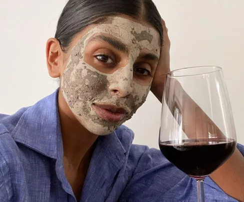 Person with a clay face mask and a blue shirt, holding a glass of red wine.