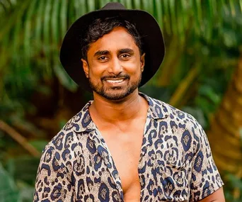 Man smiling in a leopard print shirt and black hat, standing outdoors with greenery in the background.