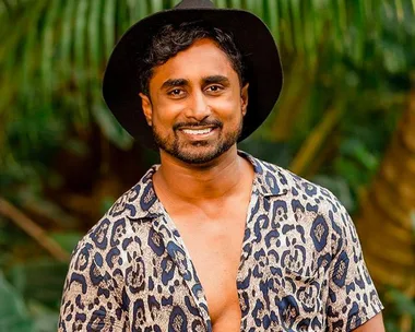 Man smiling in a leopard print shirt and black hat, standing outdoors with greenery in the background.