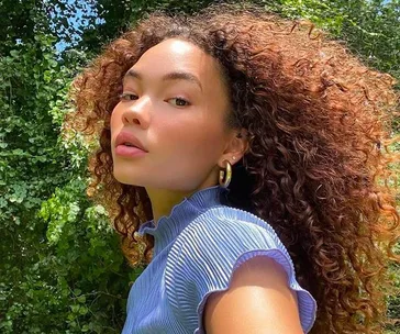 A person with curly hair wearing a blue pleated top poses outdoors with greenery in the background.