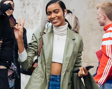 Smiling woman in green jacket and white turtleneck holds phone, giving a peace sign.