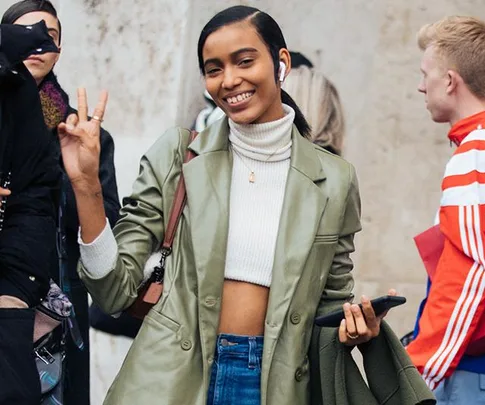 Smiling woman in green jacket and white turtleneck holds phone, giving a peace sign.