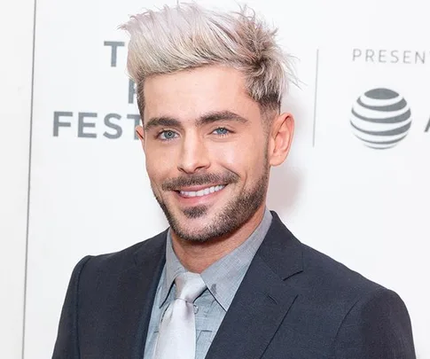 A person with short blond hair, dressed in a suit and tie, smiling at an event with a logo in the background.