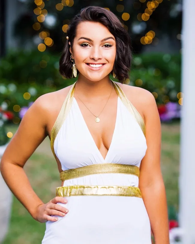 Young woman in a white and gold dress smiling, standing outdoors with blurred lights in the background.
