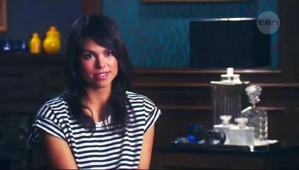 Woman with dark hair in a striped shirt sits in a room with decorative bottles and blue wallpaper.