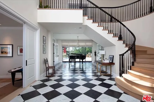 Stylish foyer with curved staircase, checkered floor, and grand piano by large windows leading to an outdoor view.