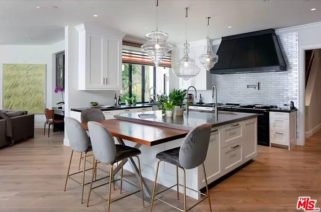 Modern kitchen with a large island, pendant lights, gray barstools, white cabinets, and black range hood.