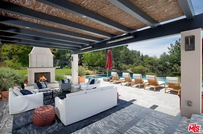 Outdoor patio with white sofas, fireplace, and poolside loungers under a pergola, surrounded by lush greenery.