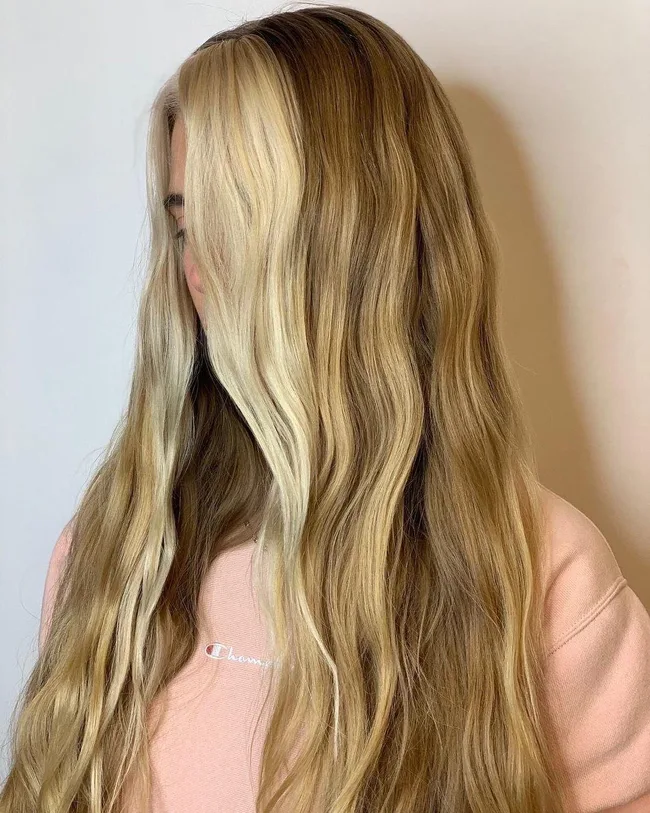 A person with long, wavy blonde and brown hair, wearing a pink sweatshirt, looking down.