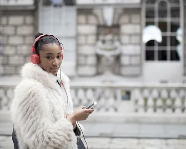 A woman in a white fur coat listens to headphones while holding a smartphone, standing outside a building.