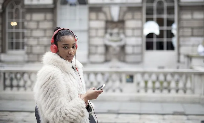 A woman in a white fur coat listens to headphones while holding a smartphone, standing outside a building.