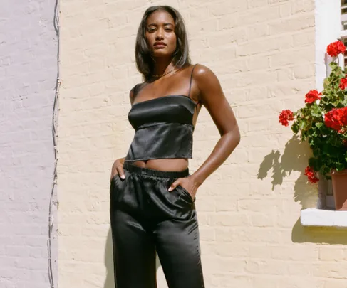 A woman in a black silk top and pants poses by a sunny wall with red flowers.