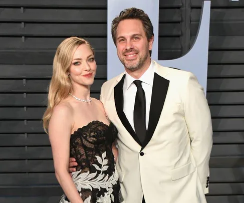 A couple poses at an event; she wears a black lace dress, he wears a white tuxedo with a black tie.