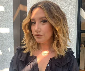 Woman with wavy blonde hair poses in a black top against a light wall.