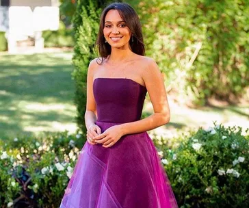 Woman in a strapless purple gown standing in a garden, smiling with hands clasped.