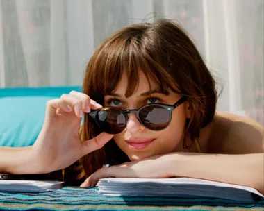 Woman with sunglasses lying on a towel, looking over papers, against a soft curtain backdrop.